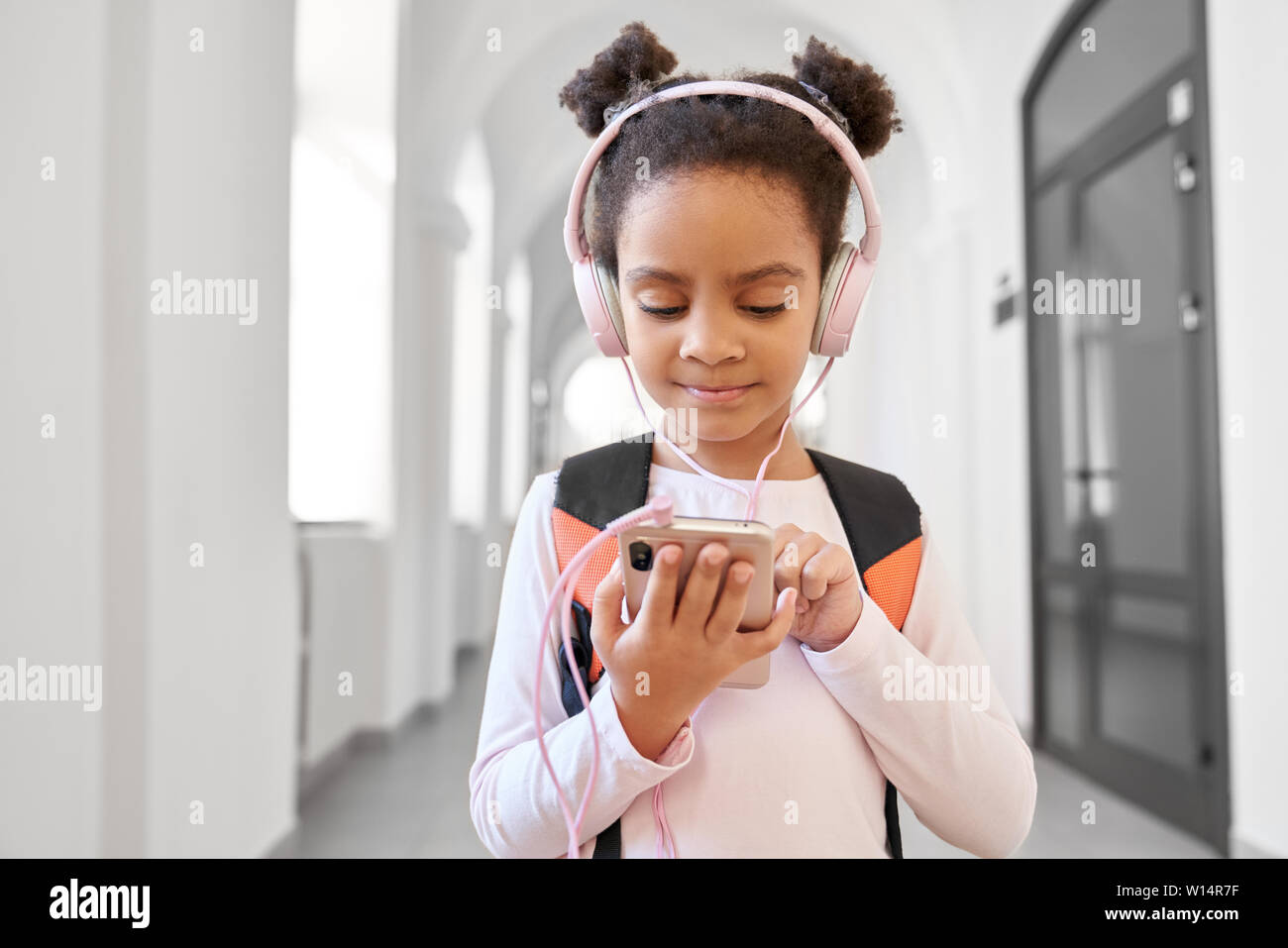 Pretty little African school girl wearing big headphones standing at long corridor and using call phone. Funny girl with backpack studying in international school. Stock Photo