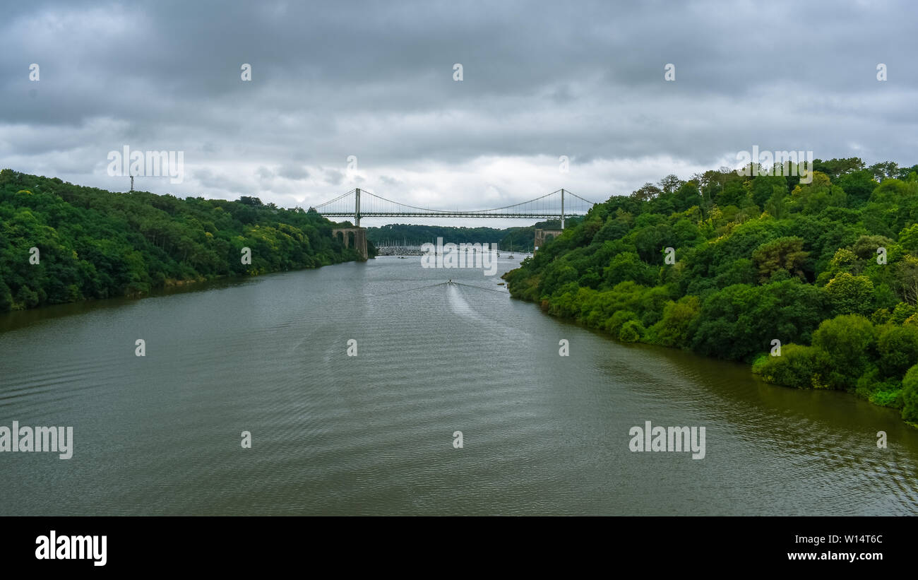 Cable-stayed bridge in a green natural forest, with a dramatic cloudy sky. Motorboat sailing on the river Stock Photo