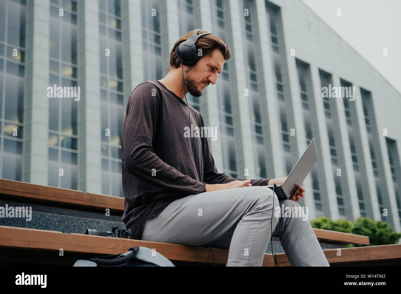 The freelance guy listens to music on headphones, backpac and works on a laptop in the center of a big city. Stock Photo