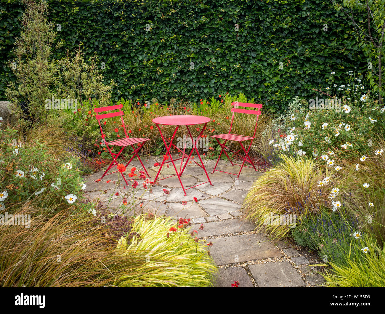 Secluded area of garden with small patio and two red chairs with metal table surrounded by summer flowers and grasses. Stock Photo