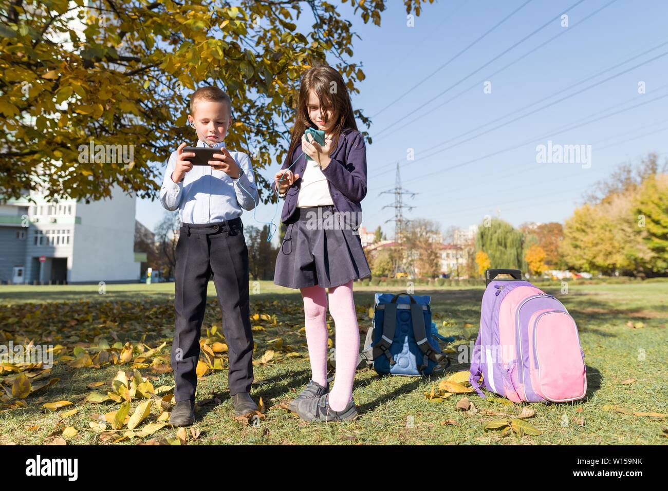 Two children 7, 8 years old with mobile phones, schoolchildren with backpacks looking into smartphones, outdoor background. Education, friendship, tec Stock Photo