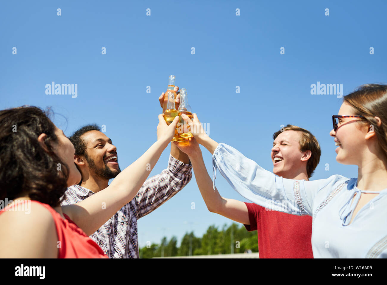 Waist up portrait of happy young friends clinking beer bottles against blue sky in Summer, copy space Stock Photo