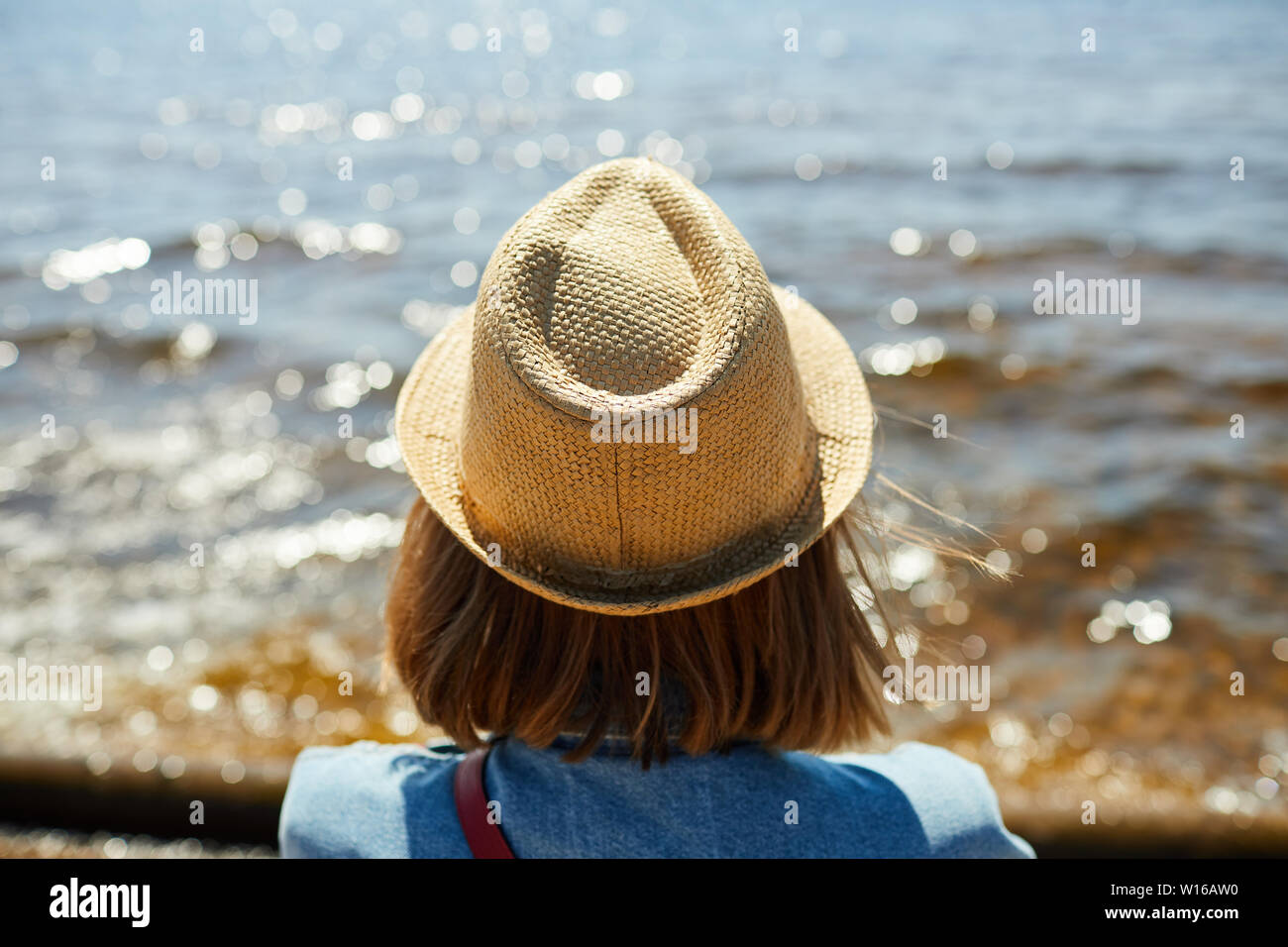 Back view portrait of young woman wearing straw hat looking out at sea in Summer, copy space Stock Photo