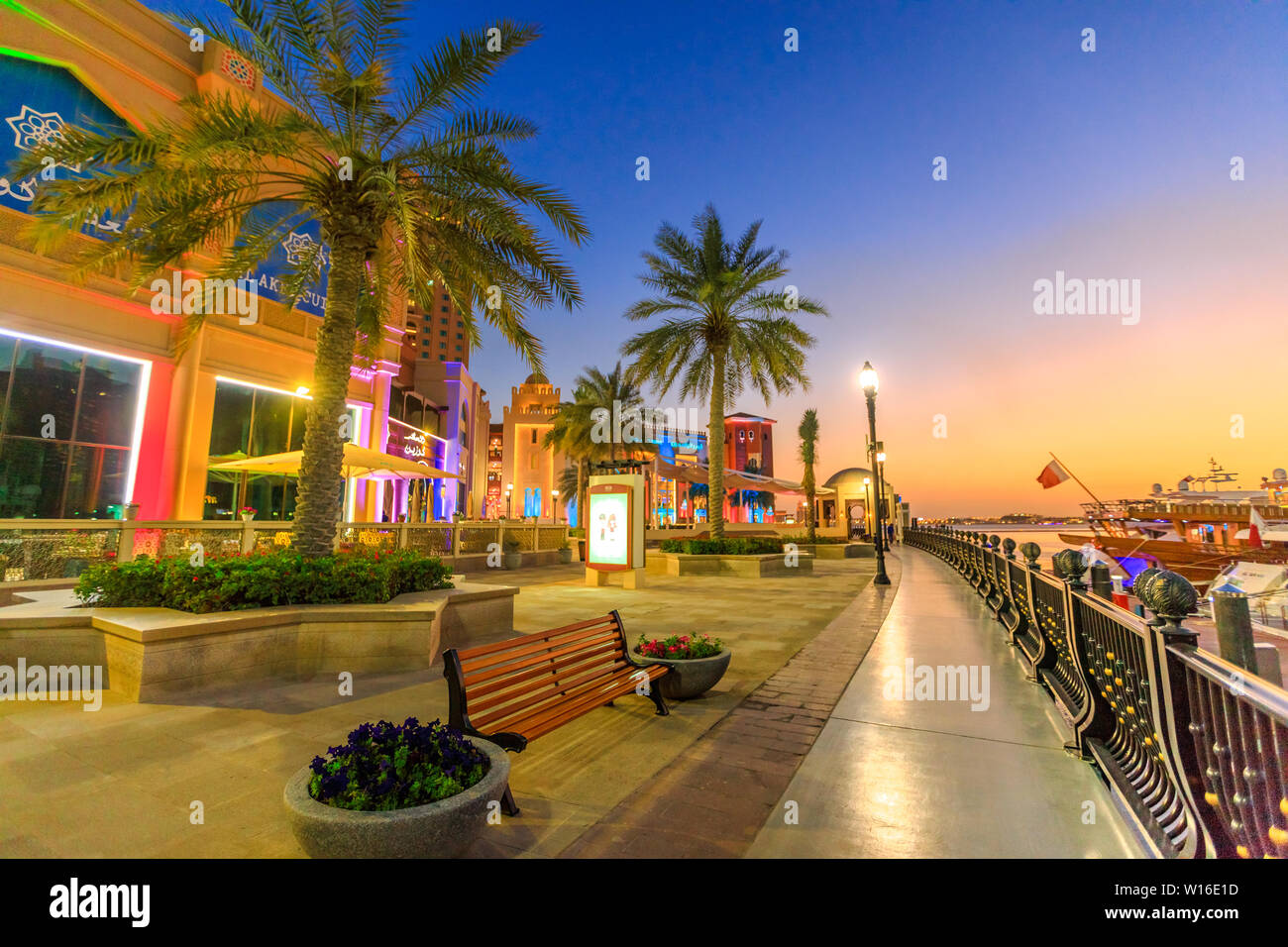 Doha, Qatar - February 18, 2019:Palm trees in walkway of Porto Arabia at the Pearl-Qatar, Doha, with skyscrapers of West Bay skyline at sunset sky Stock Photo