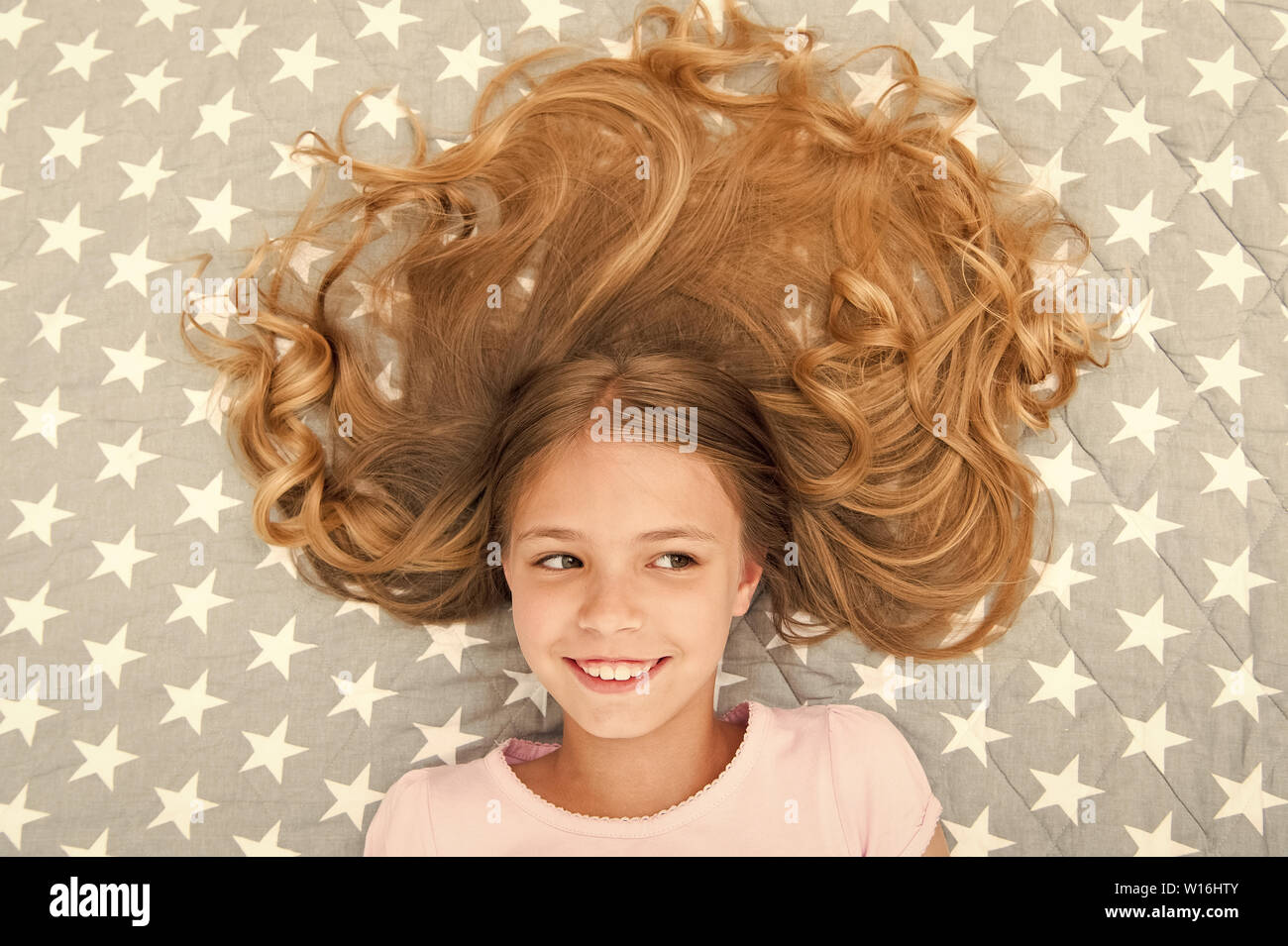 Child curly hairstyle relaxing. Conditioner mask organic oil keep hair shiny and healthy. Amazing hair tips. Keep hair curly even next morning. Girl child with long hair lay on bed top view Child curly hairstyle relaxing. Conditioner mask organic oil keep hair shiny and healthy. Amazing hair tips. Keep hair curly even next morning. Girl child with long hair lay on bed top view
