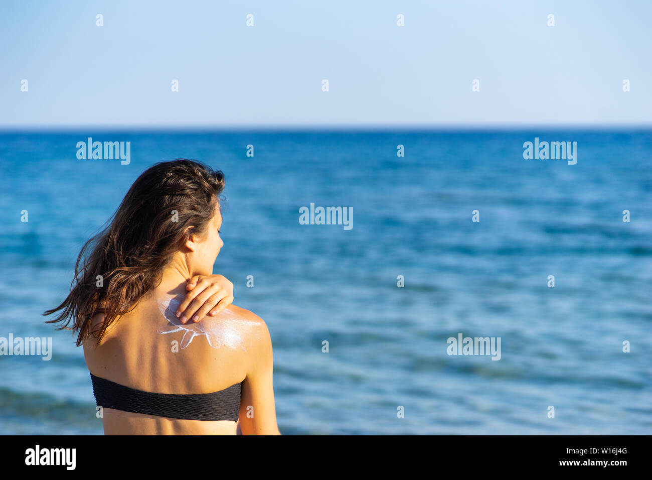 young woman with dark hair applying sun cream to her back while sitting on the beach Stock Photo