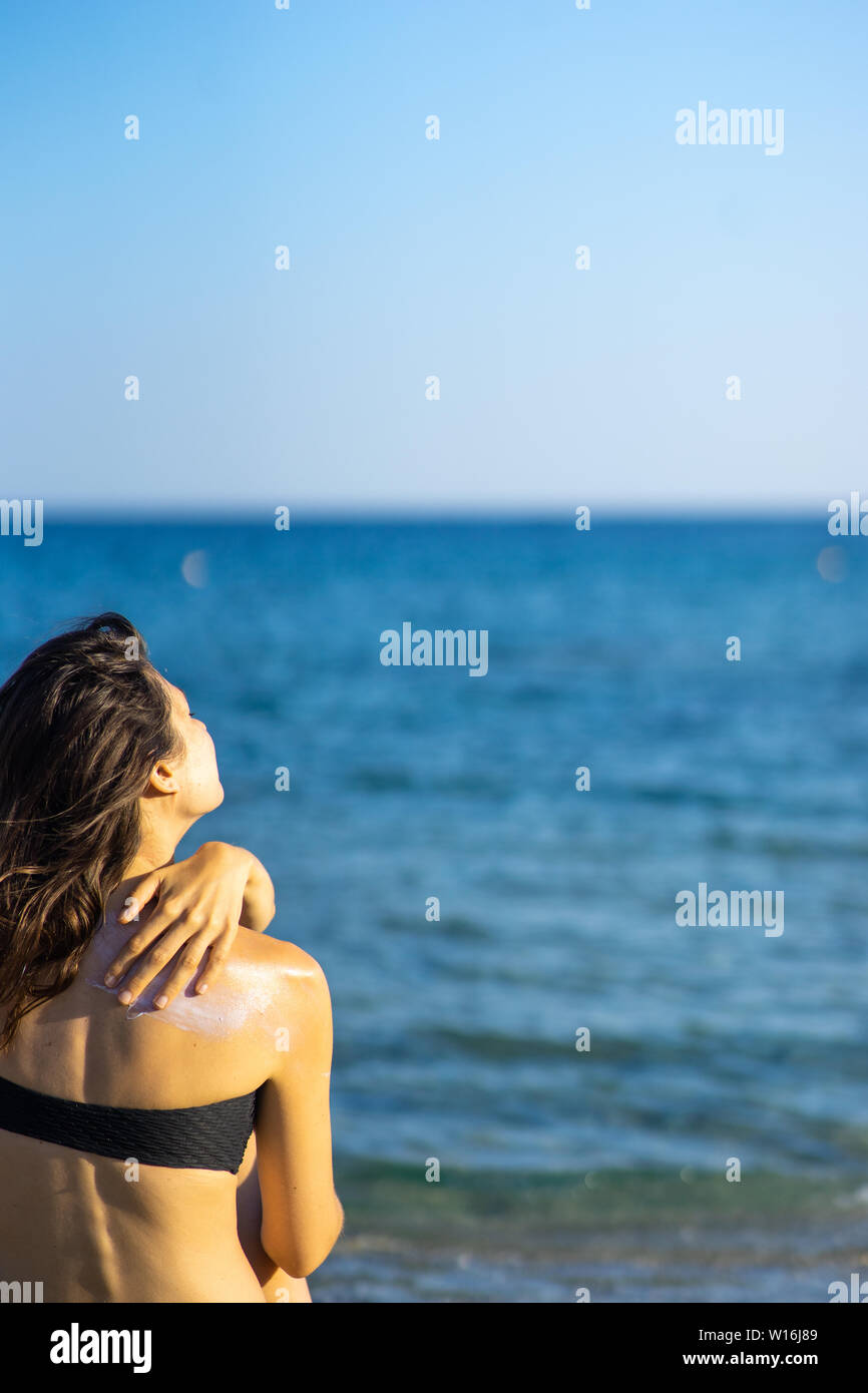 young woman with dark hair applying sun cream to her back while sitting on the beach Stock Photo