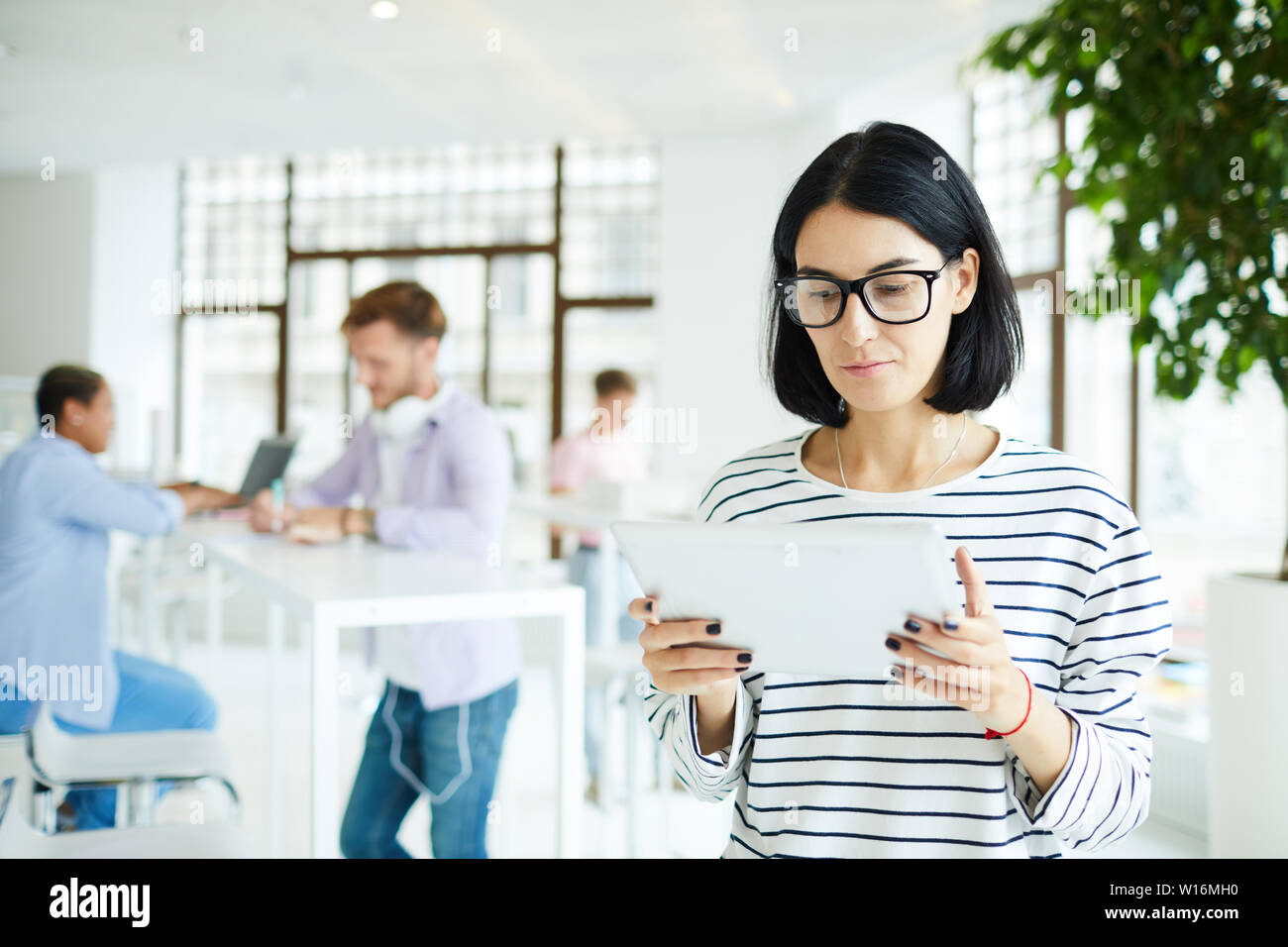 Serious pensive young female freelancer in eyeglasses standing in modern room and analyzing online sources on tablet Stock Photo