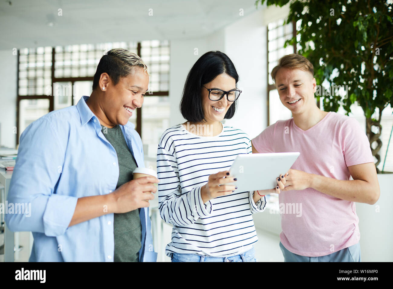 Cheerful excited young multi-ethnic colleagues in casual clothing standing in modern lobby and testing new software on tablet Stock Photo