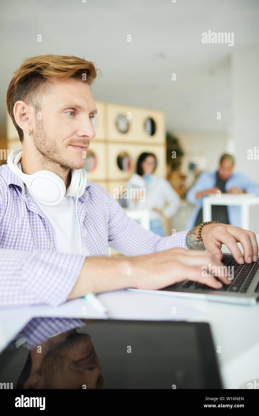 Serious focused handsome young man with headphones on neck sitting at table and using laptop while searching for online information Stock Photo