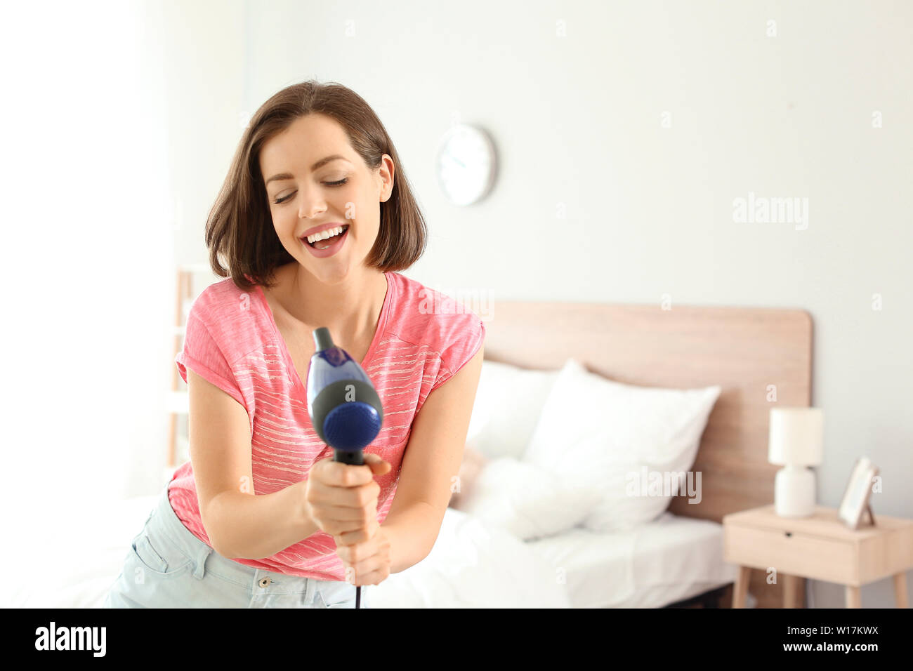 Beautiful young woman having fun with blow dryer in bedroom Stock Photo