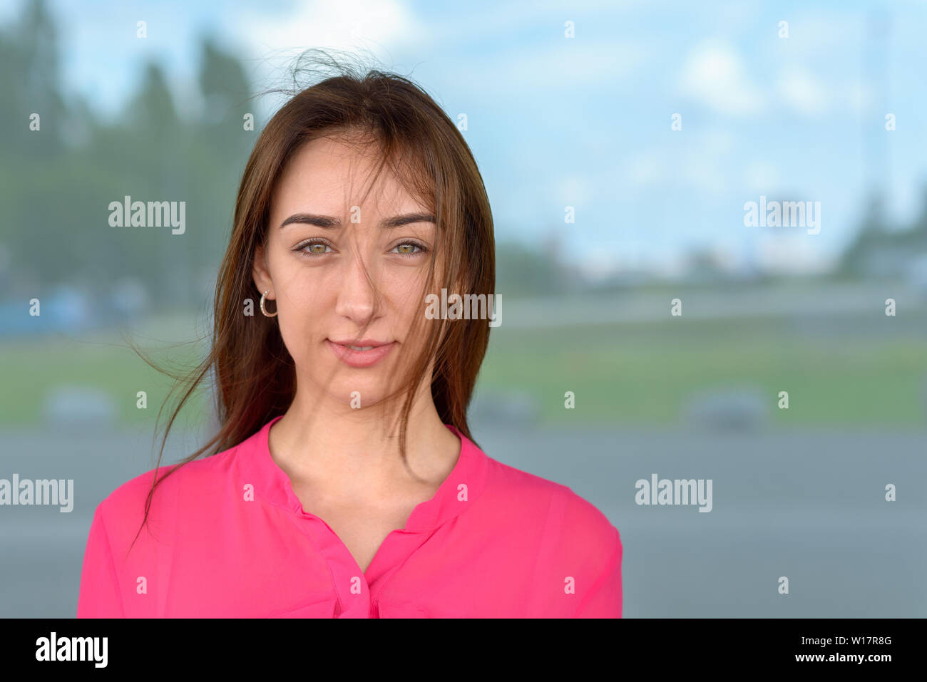 Attractive natural young woman with wisps of hair falling across her face posing in front of a large window looking at the camera with a quiet friendl Stock Photo