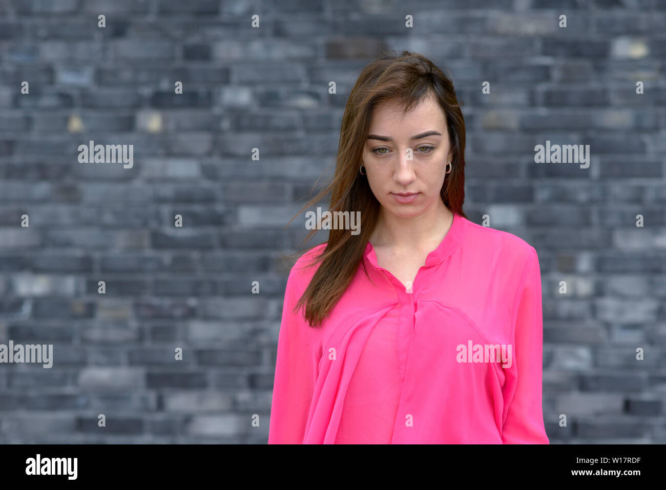 Thoughtful young woman with downcast eyes and a serious expression posing in a bright pink top in front of a grey brick wall Stock Photo