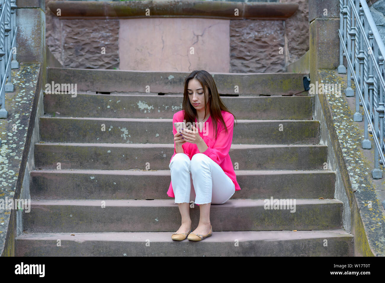 Young woman sitting on exterior cement steps using her mobile phone engrossed in reading a message Stock Photo