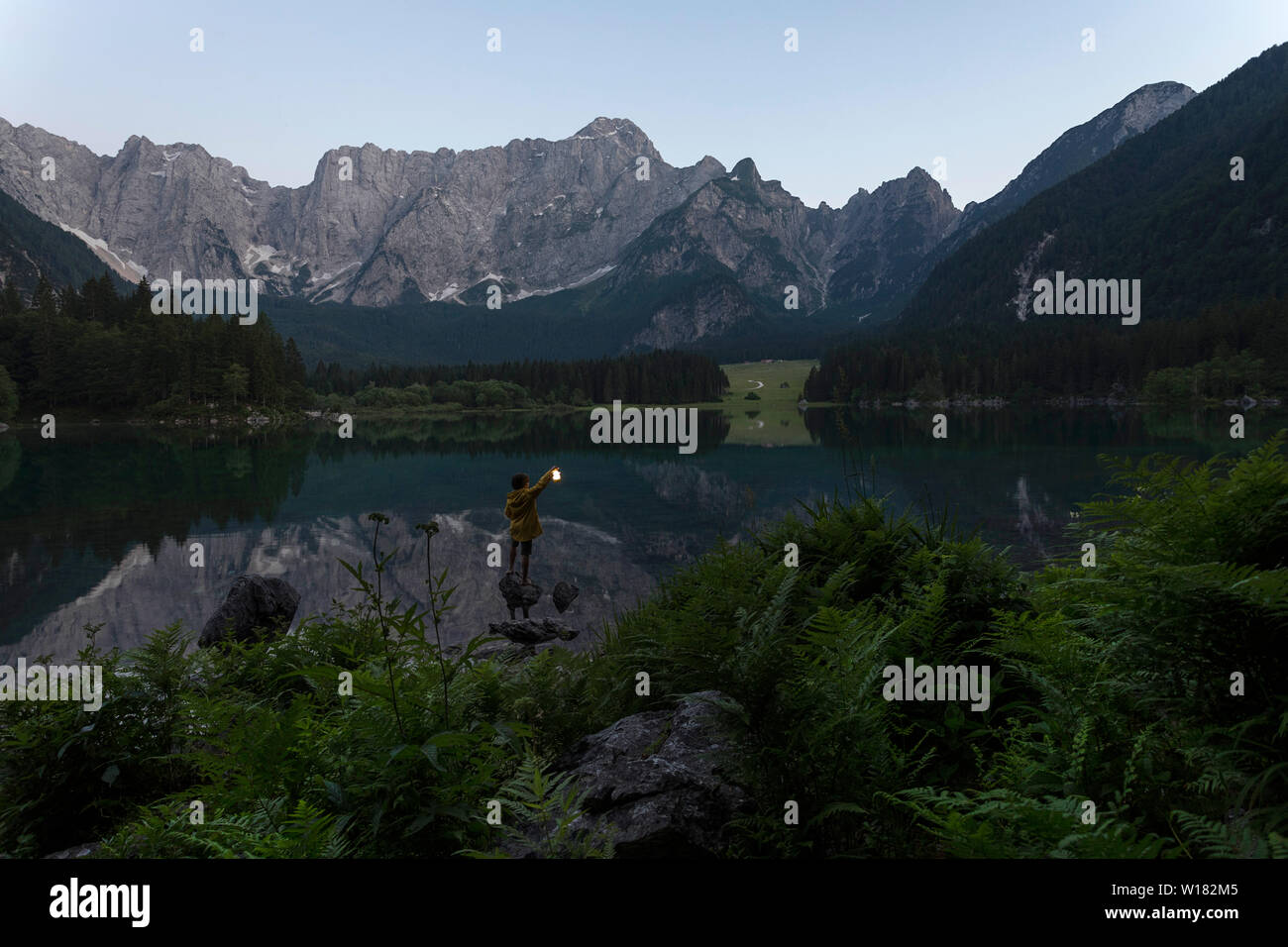 Boy in yellow rain jacket standing on the rock and holding a luminous hand lantern at scenic summer lake landscape in twilight, Laghi di Fusine, Italy Stock Photo