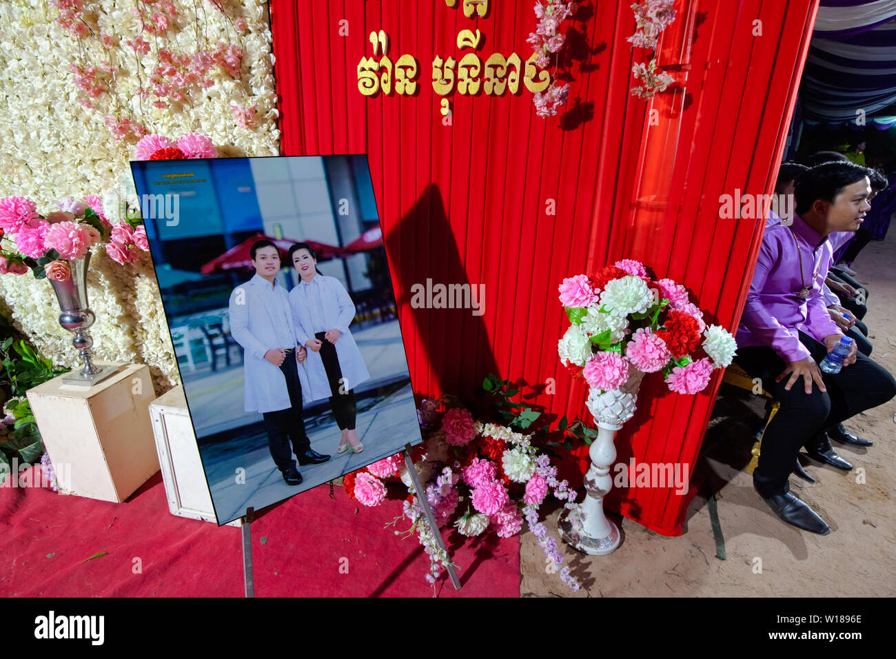 A Khmer newlywed man sits to the left of a photograph depicting him and his new wife at a traditional Khmer wedding ceremony in Siem Reap, Cambodia. Stock Photo