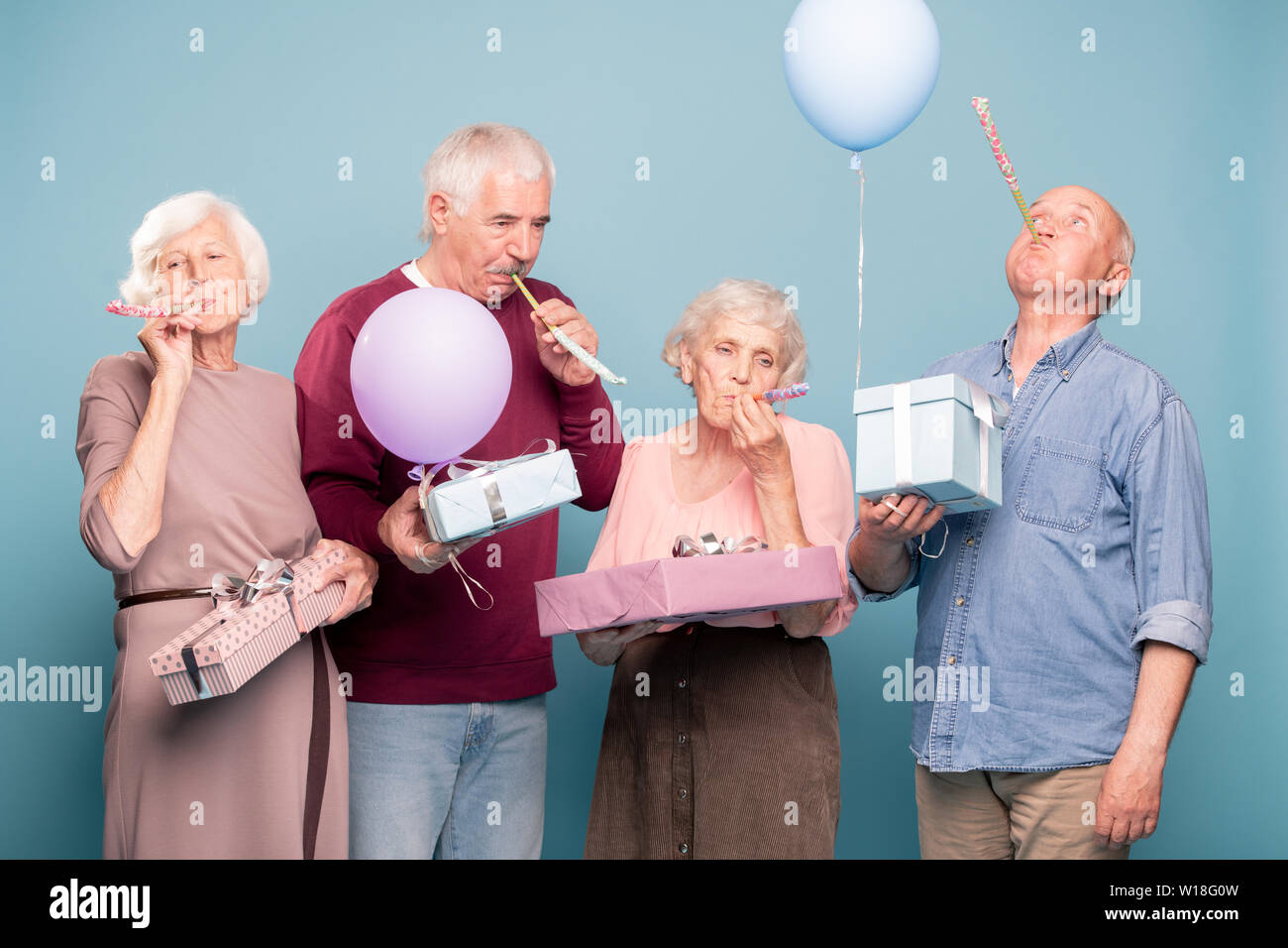 Two senior couples with giftboxes blowing whistles while having birthday fun Stock Photo