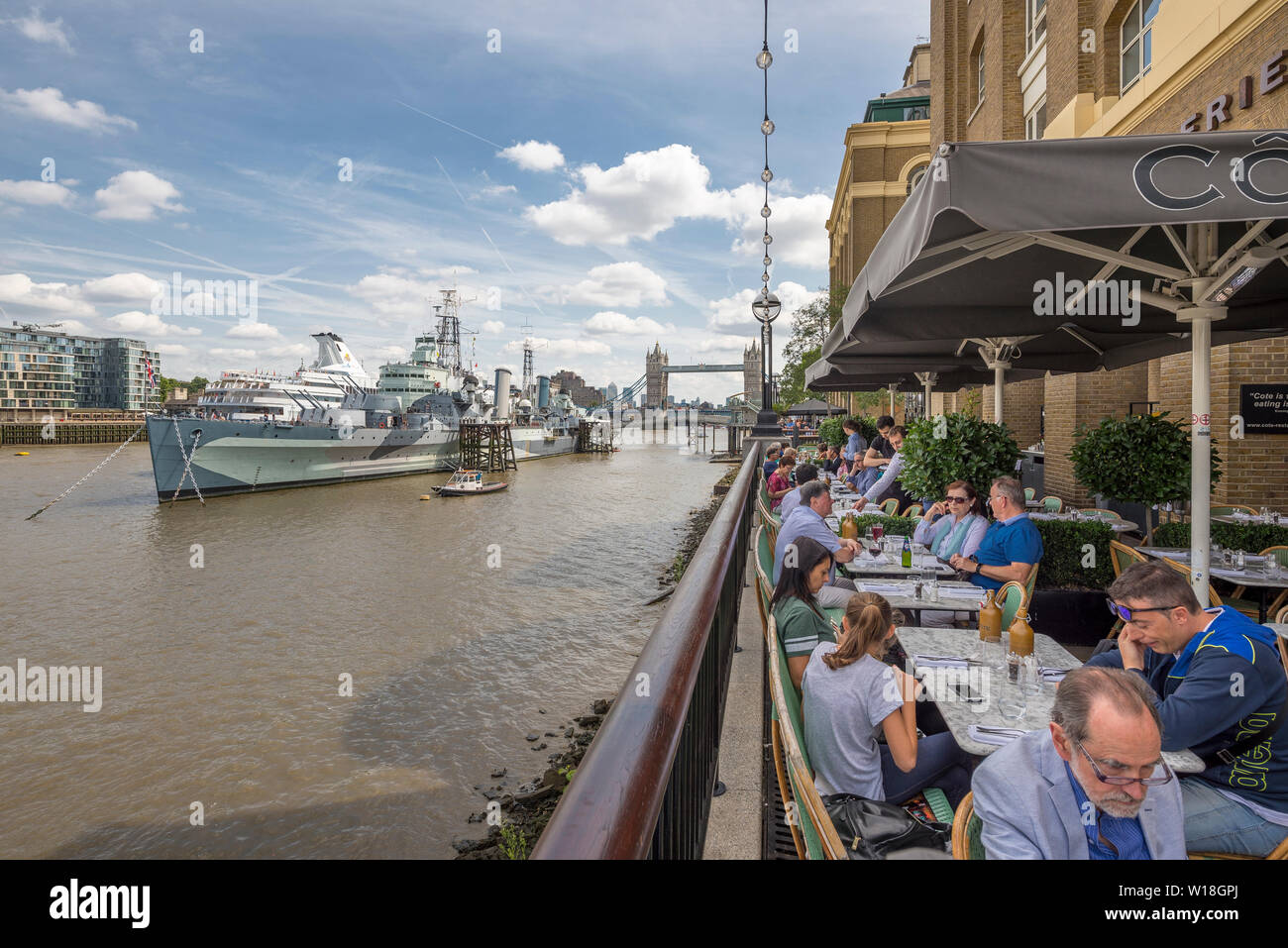 A busy restaurant terrace overlooking the river thames and HMS Belfast at the tourist hotspot area on the South Bank in Central London in summer Stock Photo