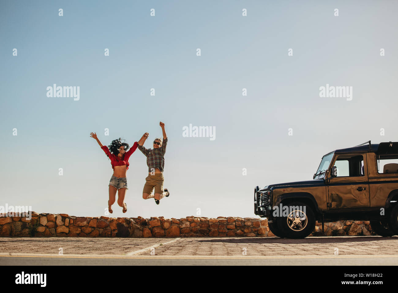 Young man and woman jumping outdoors in front of the car. Couple having a great time on road trip. Stock Photo