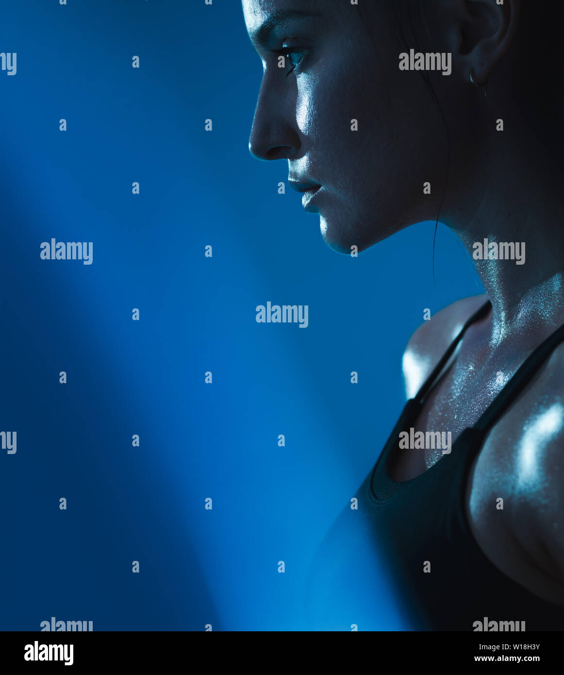 Close up of young woman in sports bra with sweat against blue background. Muscular build female relaxed after workout. Stock Photo