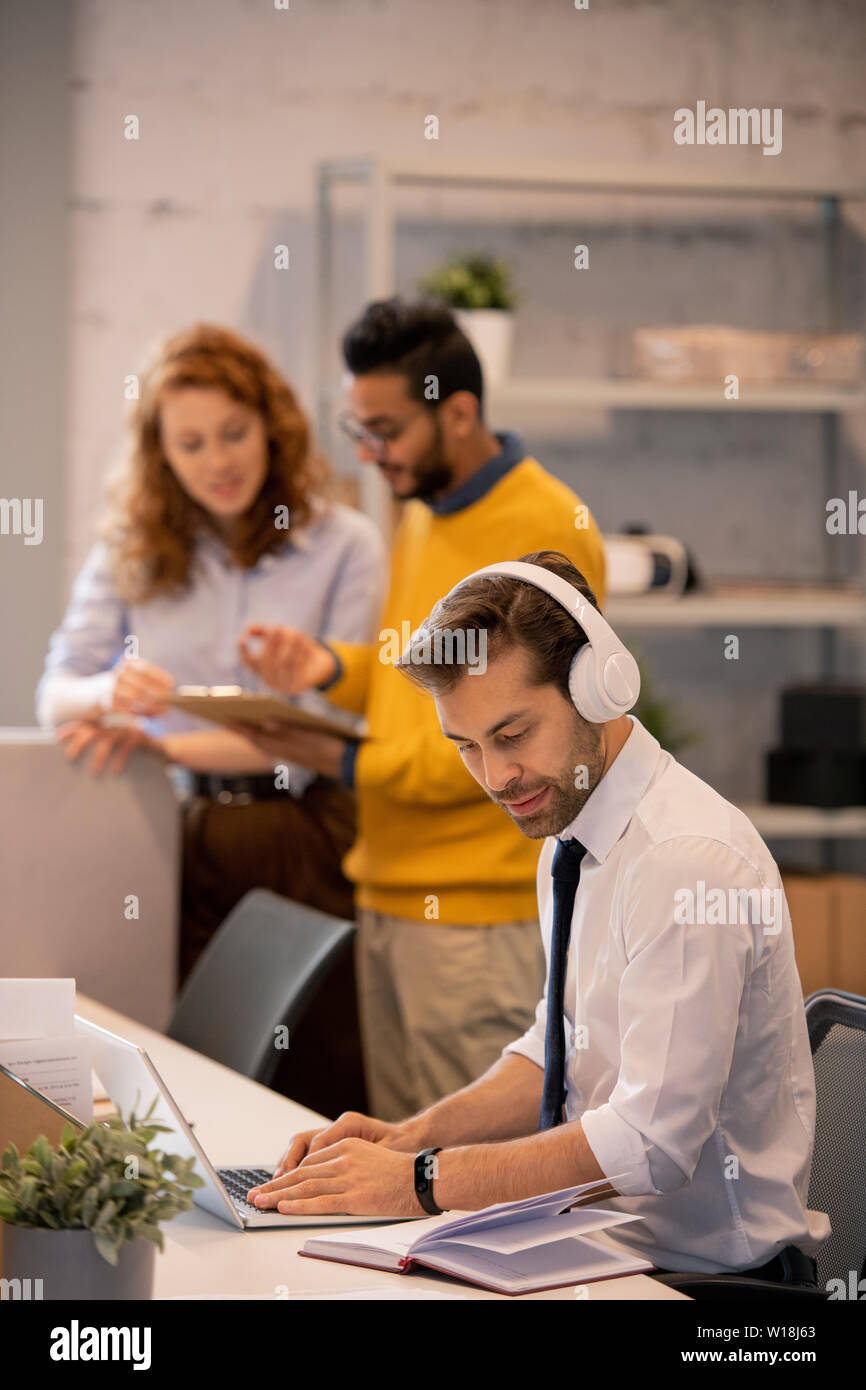 Serious concentrated young employee in headphones sitting at table and typing on laptop while adding information into presentation Stock Photo