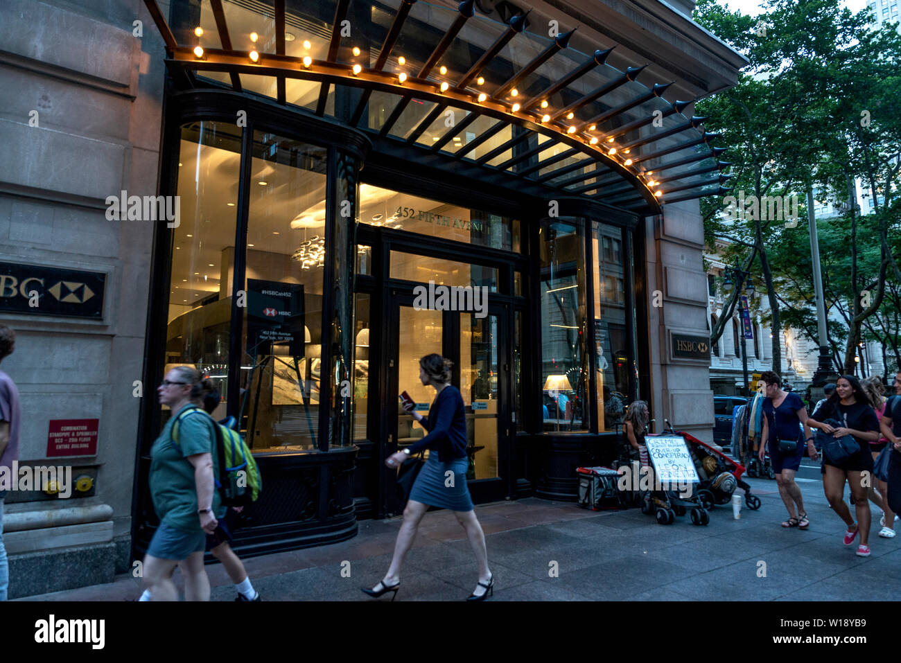 New York City, USA - August 1, 2018: Facade of a bank branch of HSBC Bank at night with a beggar and people around in 5th Avenue (Fifth Avenue), Manha Stock Photo