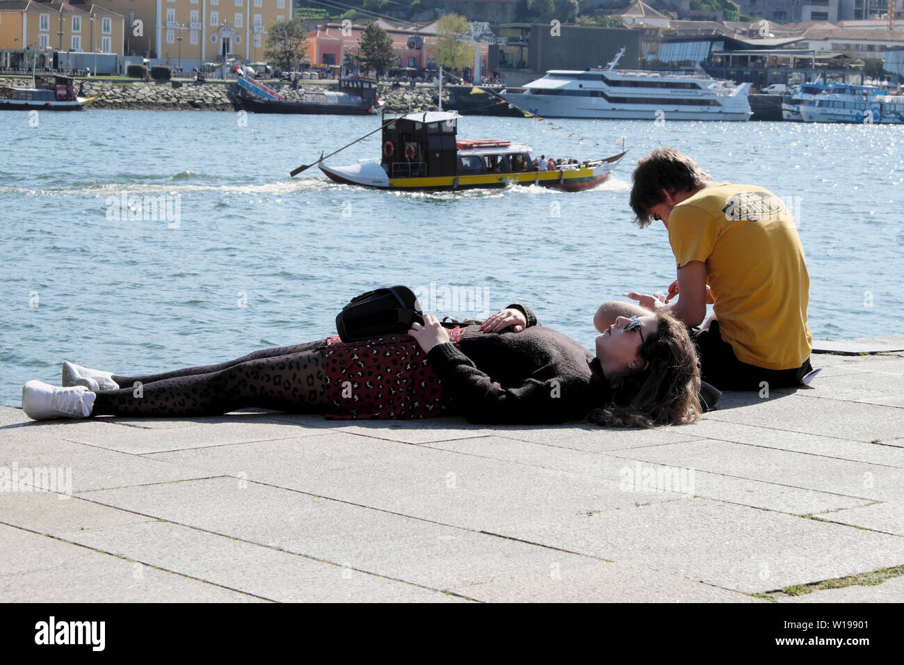 Two young people on the quay with boat passing by and woman sunbathing in spring March 2019 Ribeira Porto Oporto Portugal Europe  KATHY DEWITT Stock Photo