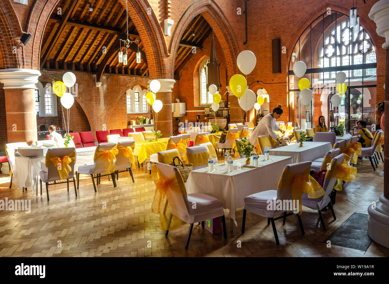 A church hall laid out with tables and chairs dressed in white fabric with yellow ribbon bows ready for a party or reception. Stock Photo