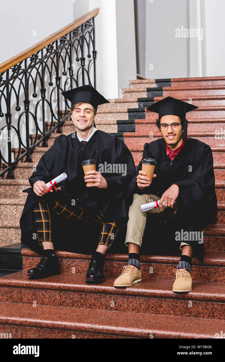 cheerful students in graduation gowns holding diplomas and paper cups ...