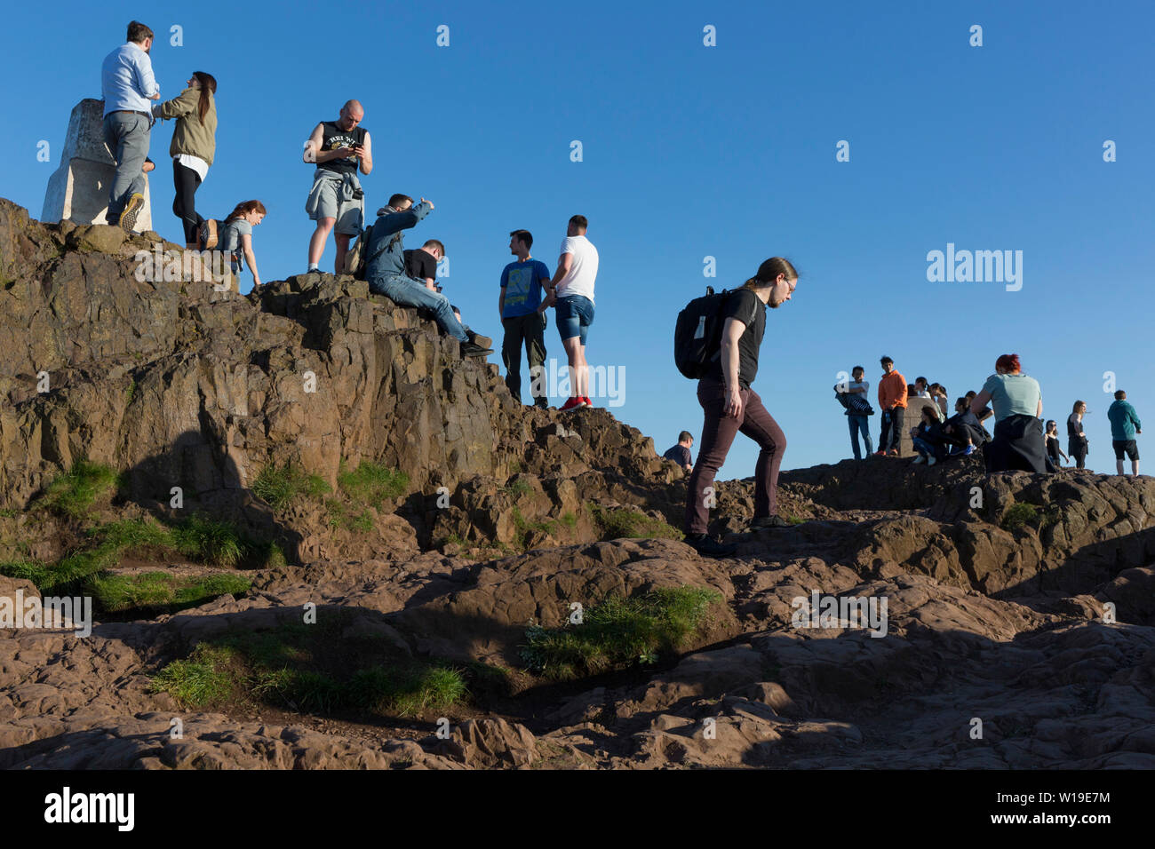 Walkers enjoy summer evening sunshine on the summit of Arthur's Seat in Holyrood Park, overlooking the city of Edinburgh, on 26th June 2019, in Edinburgh, Scotland. Arthur's Seat is an extinct volcano which is considered the main peak of the group of hills in Edinburgh, Scotland, which form most of Holyrood Park, described by Robert Louis Stevenson as 'a hill for magnitude, a mountain in virtue of its bold design'. The hill rises above the city to a height of 250.5 m (822 ft), providing excellent panoramic views of the city and beyond. Stock Photo