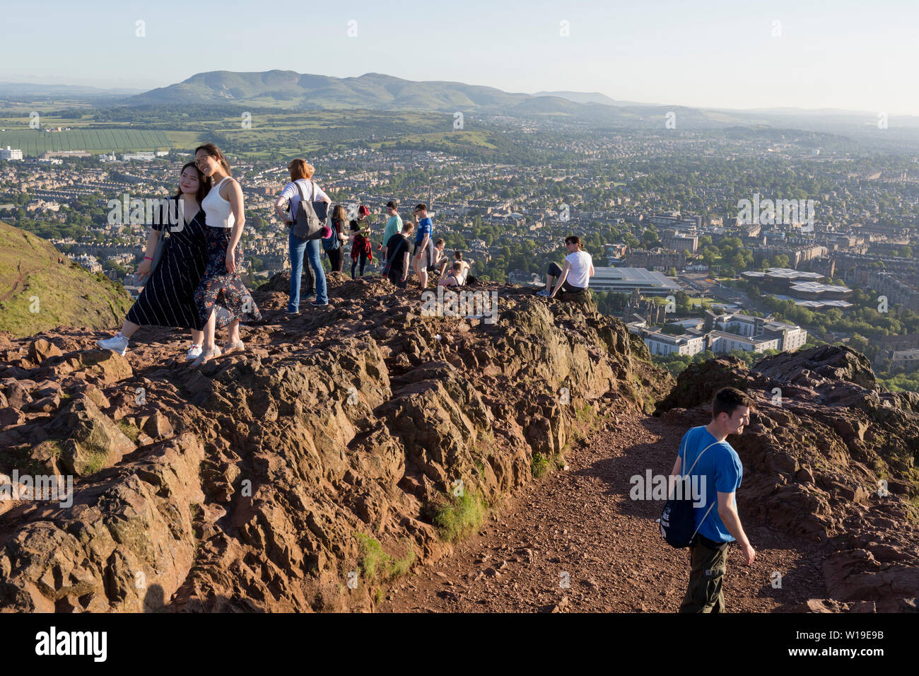 Walkers enjoy summer evening sunshine on the summit of Arthur's Seat in Holyrood Park, overlooking the city of Edinburgh, on 26th June 2019, in Edinburgh, Scotland. Arthur's Seat is an extinct volcano which is considered the main peak of the group of hills in Edinburgh, Scotland, which form most of Holyrood Park, described by Robert Louis Stevenson as 'a hill for magnitude, a mountain in virtue of its bold design'. The hill rises above the city to a height of 250.5 m (822 ft), providing excellent panoramic views of the city and beyond. Stock Photo
