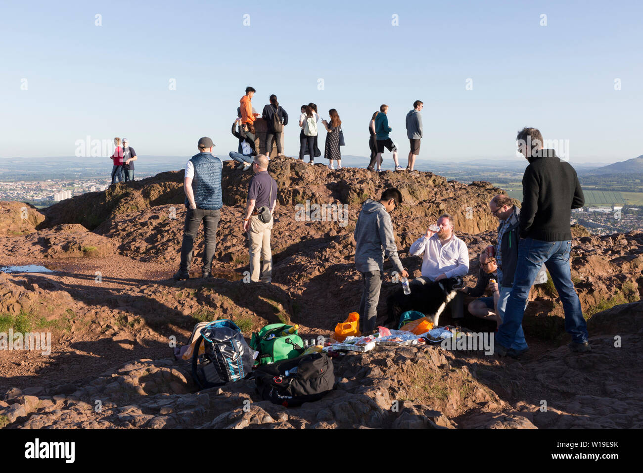 Visitors and a group of men with drinks and food enjoy summer evening sunshine on the summit of Arthur's Seat in Holyrood Park, overlooking the city of Edinburgh, on 26th June 2019, in Edinburgh, Scotland. Arthur's Seat is an extinct volcano which is considered the main peak of the group of hills in Edinburgh, Scotland, which form most of Holyrood Park, described by Robert Louis Stevenson as 'a hill for magnitude, a mountain in virtue of its bold design'. The hill rises above the city to a height of 250.5 m (822 ft), providing excellent panoramic views of the city and beyond. Stock Photo