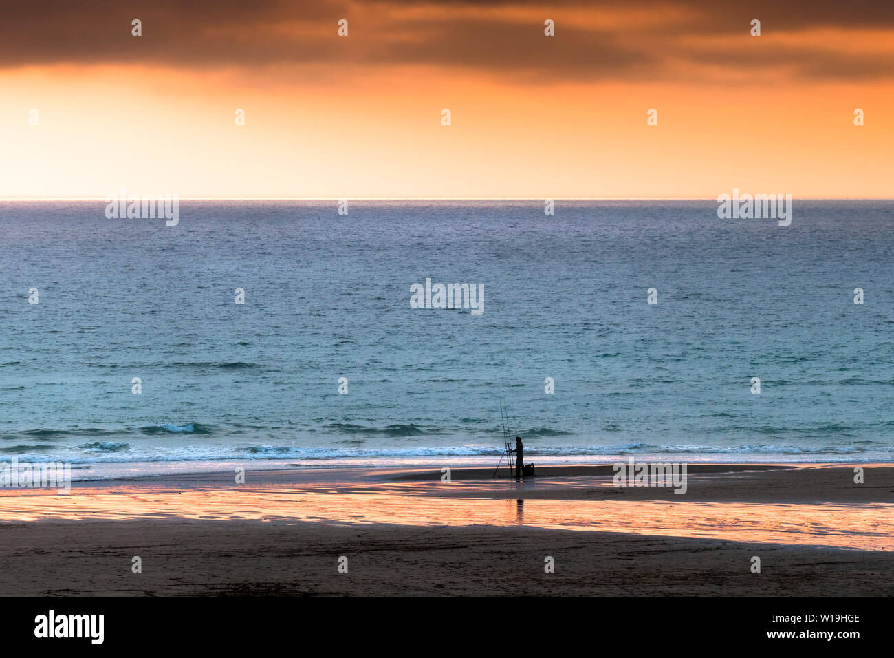 A lone angler fishing from the shore during an intense sunset over Fistral in Newquay in Cornwall. Stock Photo