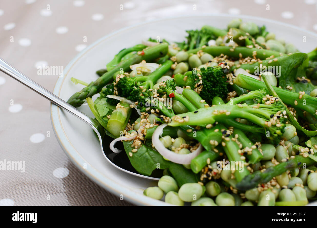 Serving spoon in a green spring salad of broccolini, broad beans, shallots and green beans, sprinkled with nigella and sesame seeds Stock Photo