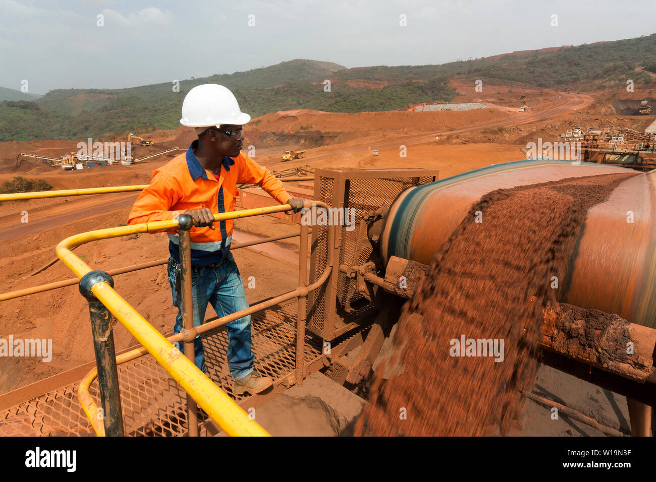 Mining operations for transporting & managing iron ore. Fines ore on conveyor belt to stacker and operator overseeing operation with mine behind. Stock Photo