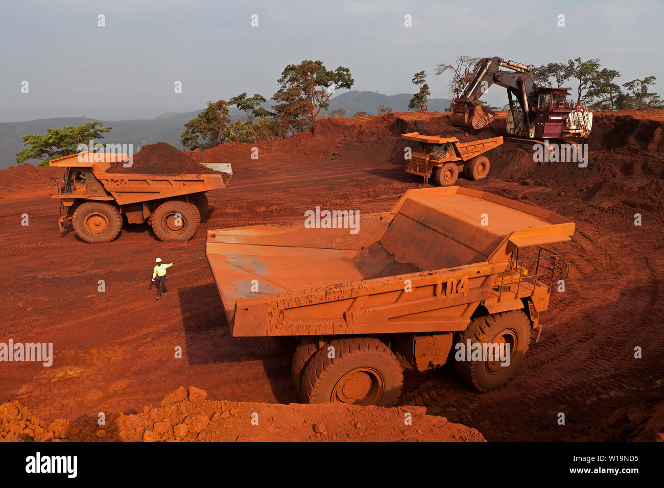 Mining operations for transporting & managing iron ore. Haul trucks being loaded with high grade haematite by excavator on bench of surface mine pit. Stock Photo