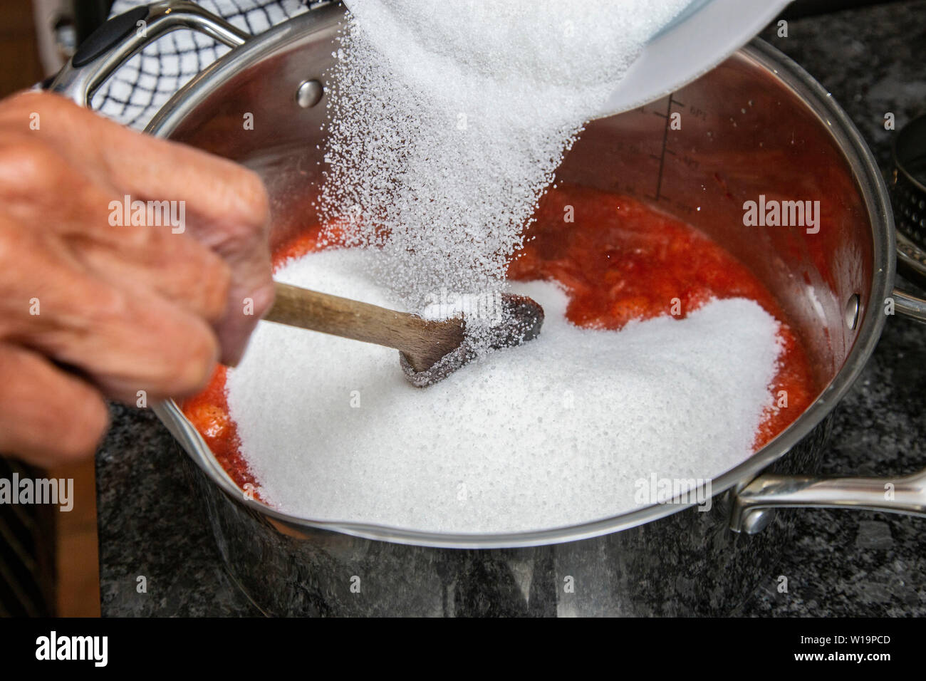 Adding sugar to strawberries as strawberry jam is made on an induction hob in a stainless steel