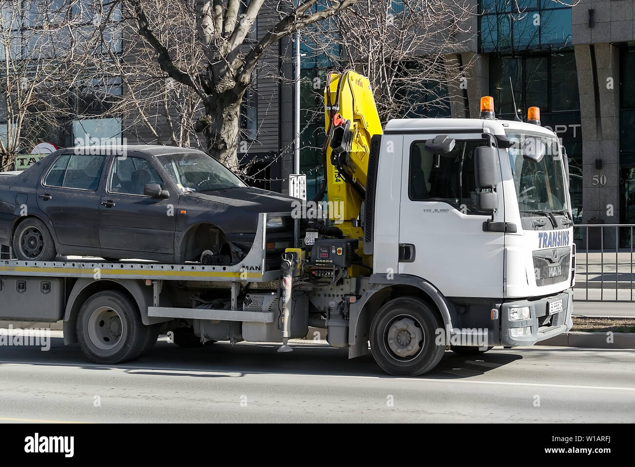 Varna, Bulgaria, February 28, 2019. A tow truck with hook and chain transports a car without a front wheel along the city street during the day. The c Stock Photo