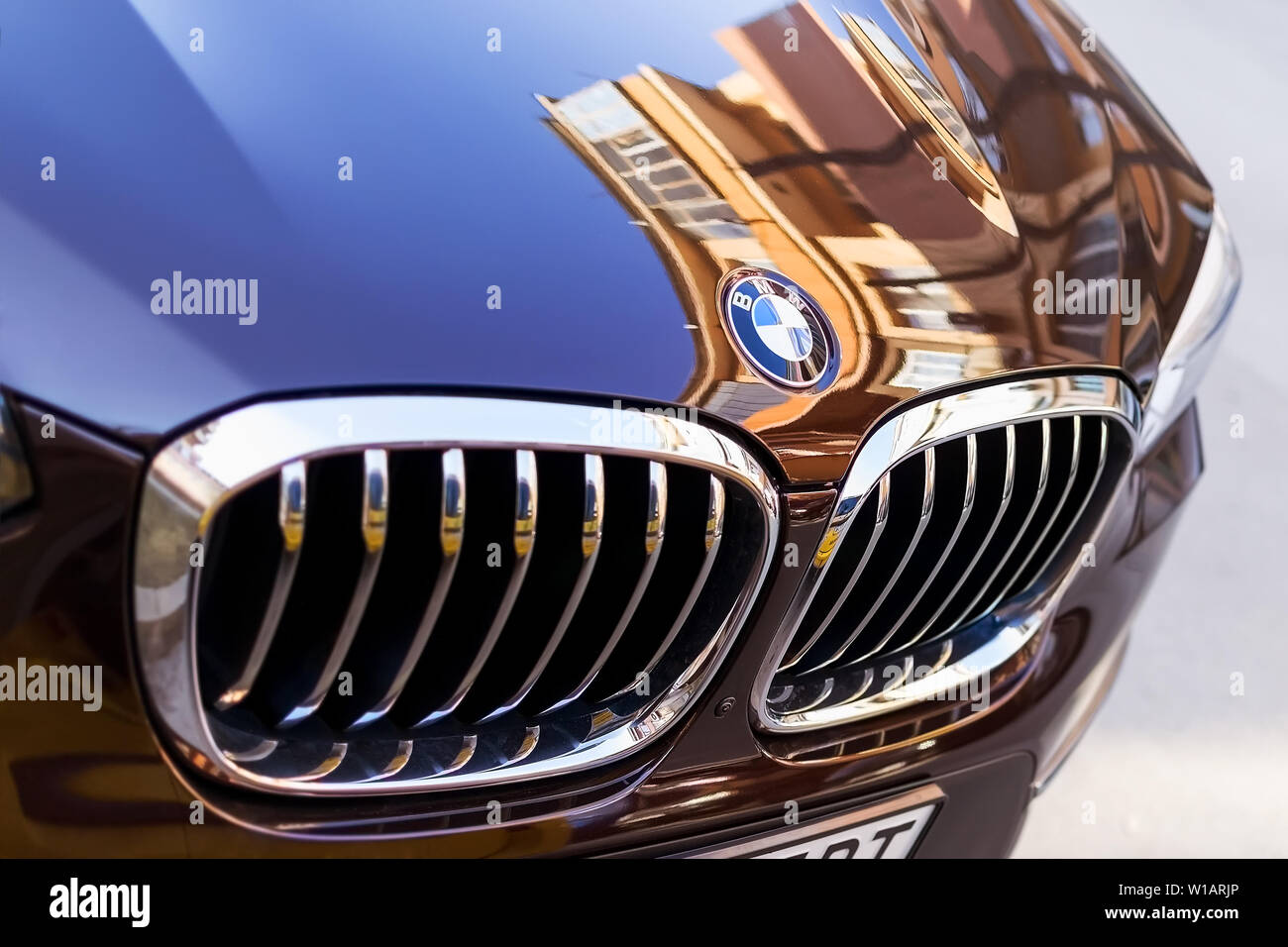 Chrome BMW logo sign close-up. Car grill and front hood of black blue BMW on an outdoor parking on a sunny day. City reflections on car hood. Stock Photo