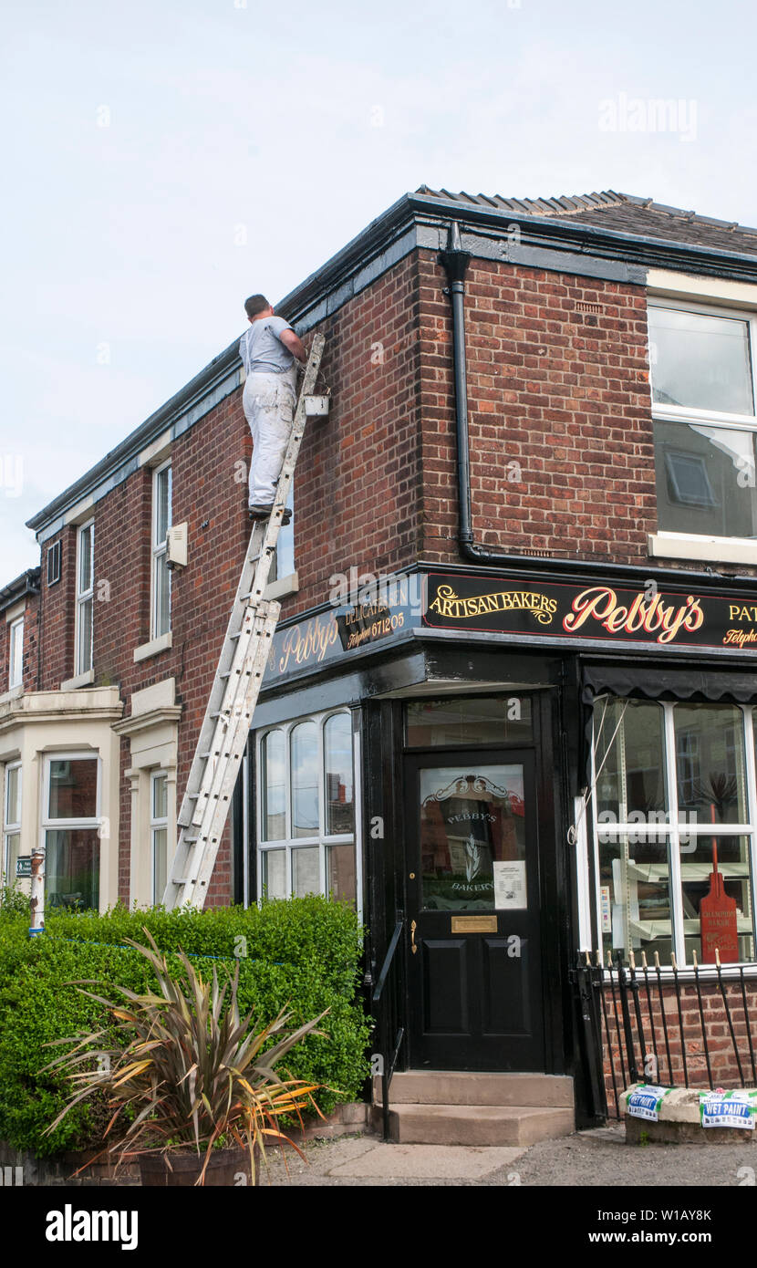 Painter on extension ladder painting black roof line and guttering above a Delicatessen Artisan bakers shop Stock Photo