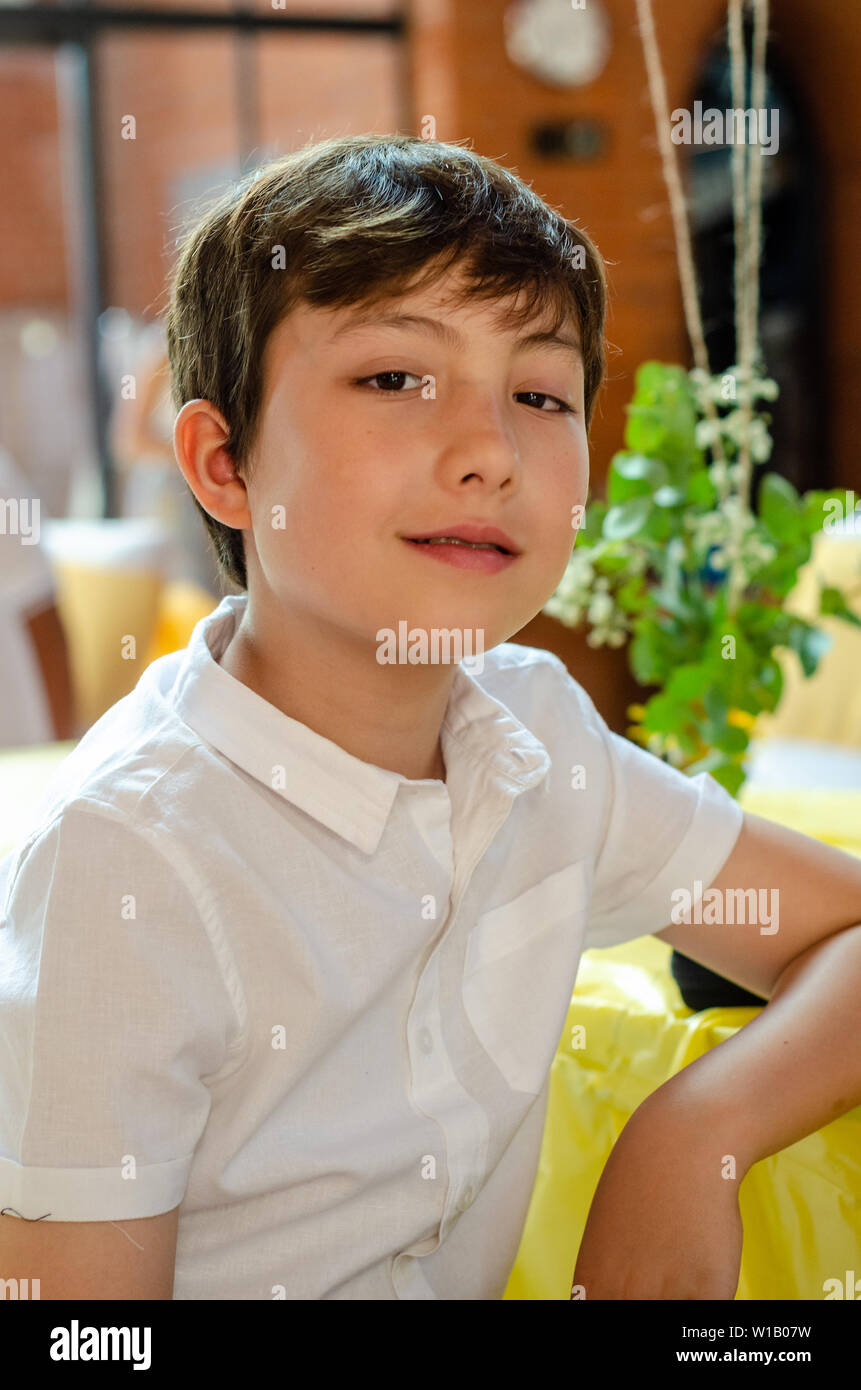 A portrait of a young boy with brown hair and eyes wearing a white short sleeved shirt looking directly into the camera. Stock Photo
