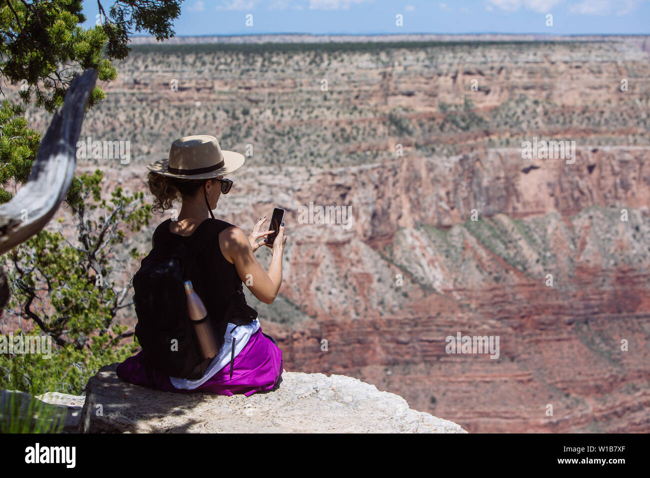 Tourist woman with hat and backpack taking a picture with a cell phone sitting on a cliff in the South Rim, Grand Canyon National Park, Arizona, USA Stock Photo