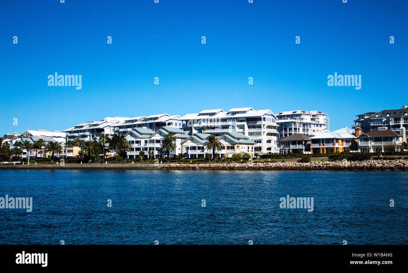Large waterside houses built in front of condominium apartments with riverside grass frontage, stone retaining wall and blue river against clear blue Stock Photo