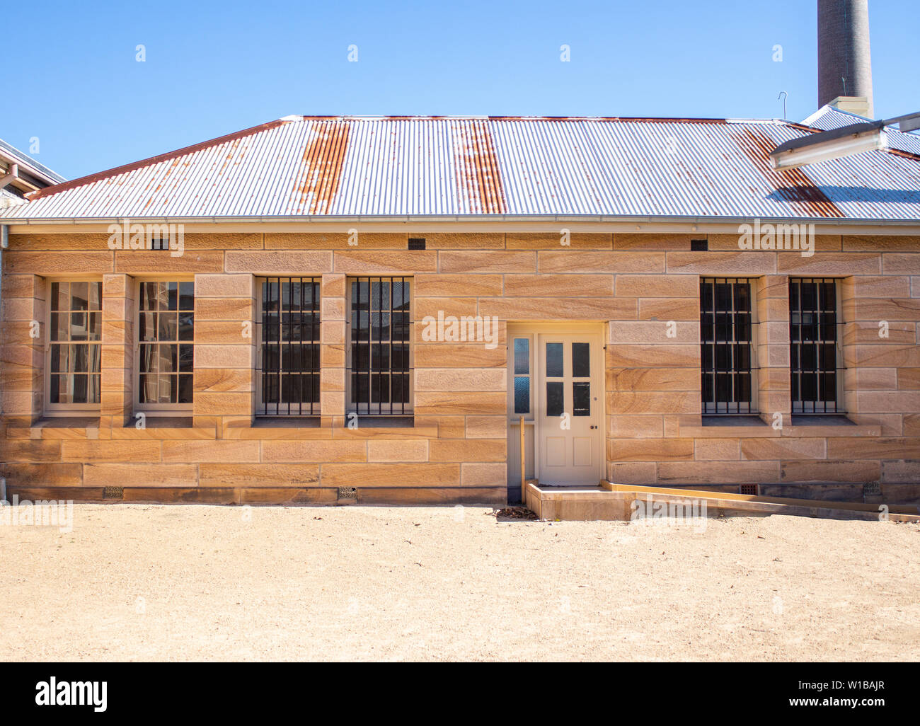 Sandstone convict brick building with corrugated iron roof, large tall windows, security grill, pebble courtyard against clear blue sky Stock Photo