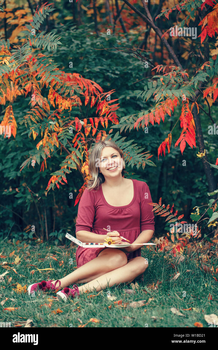 Smiling Caucasian woman artist drawing in open plein air in autumn fall park outside with pencils on paper in album. Lifestyle activity hobby. Freelan Stock Photo