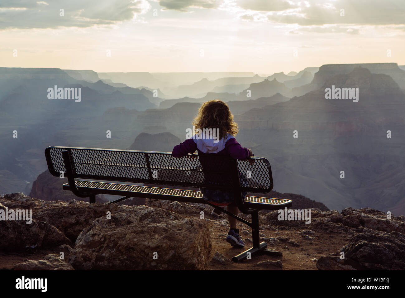 Back Caucasian, curly haired tourist woman watching sunset from a bench in Desert View, Grand Canyon National Park, Arizona, USA Stock Photo