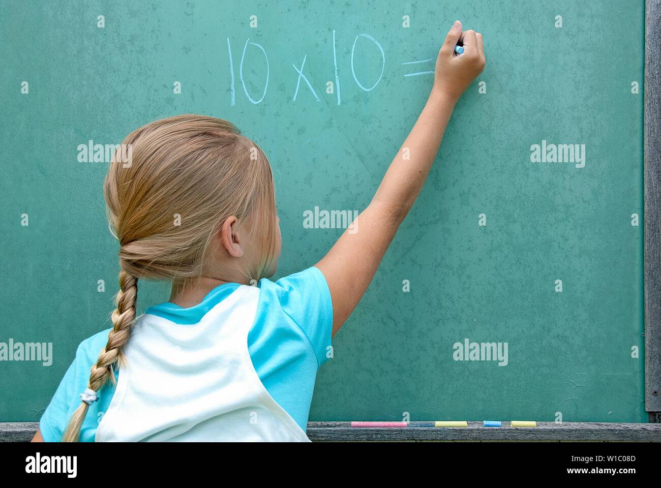 Young Caucasian girl writing math problem on green chalkboard Stock Photo