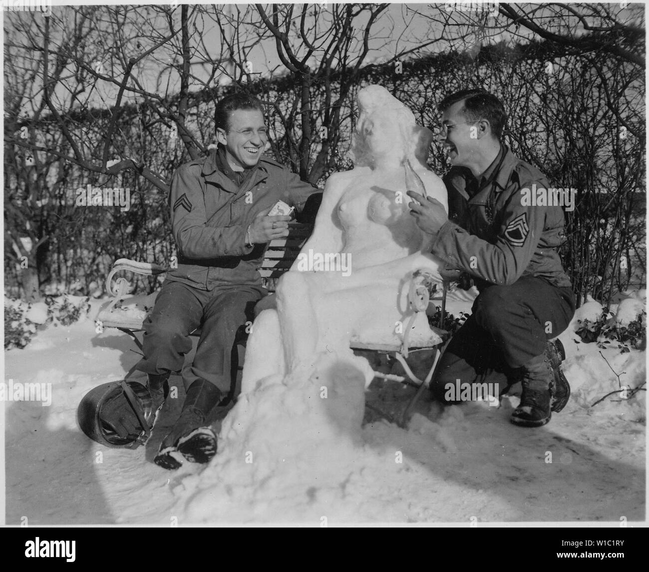 Corporal. Bernard Butnik, Cleveland Heights, Ohio, and Sergeant. Richard Goodbar, Russellville, Arkansas, offer Agnes their snow woman, cigarettes and a coke. European theater, January 14, 1945.; General notes:  Use War and Conflict Number 893 when ordering a reproduction or requesting information about this image. Stock Photo