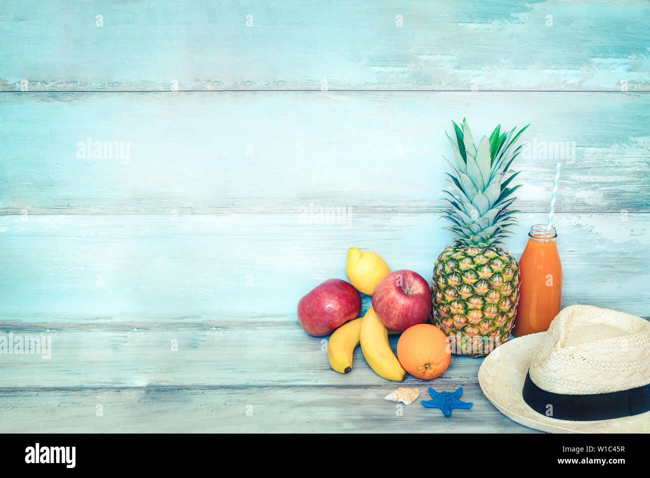 Summer concept stillife - a pile of fresh fruits, straw hat and a bottle of multivitamin juice in front of a blue rustic wooden background. Stock Photo