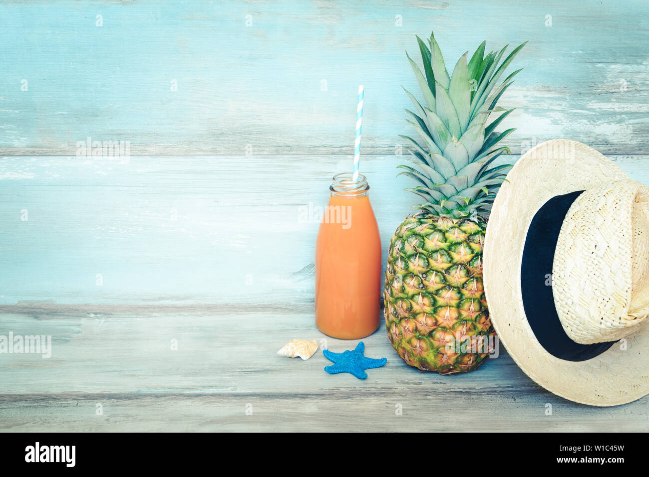 Summer concept stillife - ripe pineapple, straw hat and a bottle of multivitamin juice in front of a blue rustic wooden background. Stock Photo